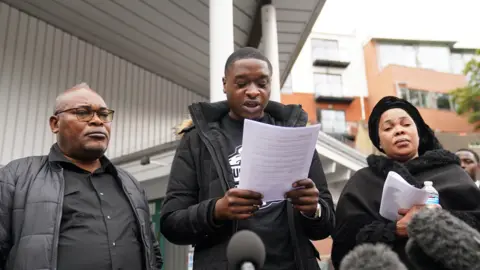 PA Media Chris Kaba's cousin Jefferson Bosela reads a statement alongside Chris's parents Prosper Kaba (left) and Helen Lumuanganu