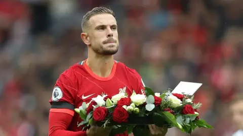 Reuters Liverpool's Jordan Henderson carrying flowers as a tribute to Andrew Devine