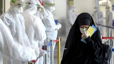 Getty Images Medical staff in protective gear distribute leaflets to Iraqi passengers returning from Iran, 5 March 2020