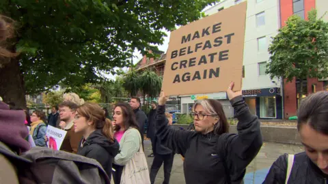 BBC A woman holds up sign that reads: MAKE BELFAST CREATE AGAIN