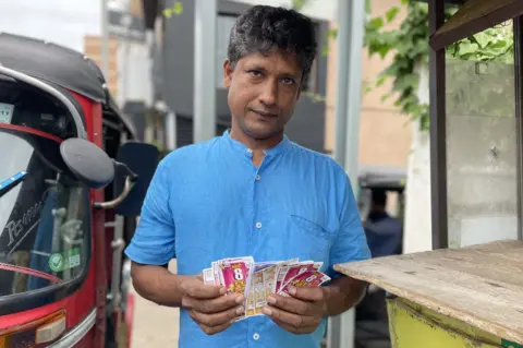 Siri, wearing a blue shirt, fans out the lottery tickets he has bought