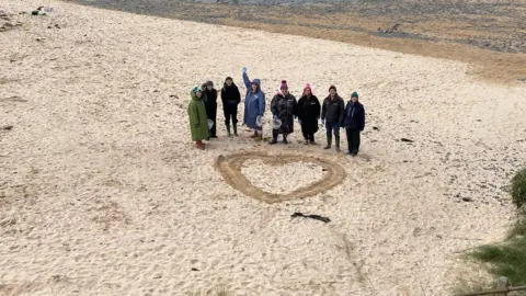 BBC People standing by heart in sand after beach clean at Seven Bays