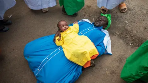 EDWIN NDEKE/GETTY IMAGES A man cradles a baby. Other people stand around in a circle.