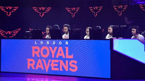 Getty Images The London Royal Ravens competing in 2020, the competitors are sat behind a blue desk which has the London Royal Ravens logo in white and red writing on a blue background, and they are looking at computer screens