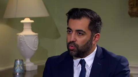 Humza Yousaf wearing a navy suit and tie sitting in a room with light green walls. A white lamp sits behind him.