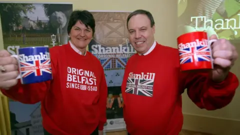 PA Media Arlene Foster and Nigel Dodds wearing Shankill-branded sweaters and holding Shankill-branded mugs in 2008