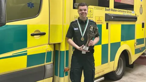 SWASFT Leon in uniform holding the World Irish Dance Champion trophy, with a South Western Ambulance Service NHS Foundation Trust vehicle.