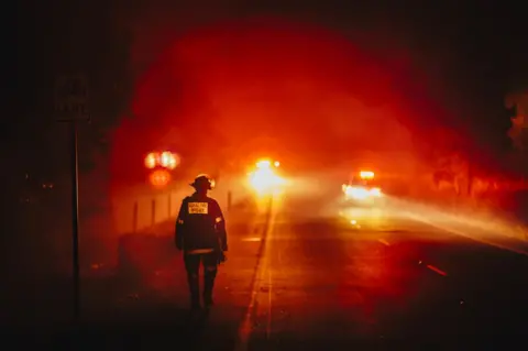 Cam Neville A firefighter walks along a roadside, framed by the red flow of distant lights