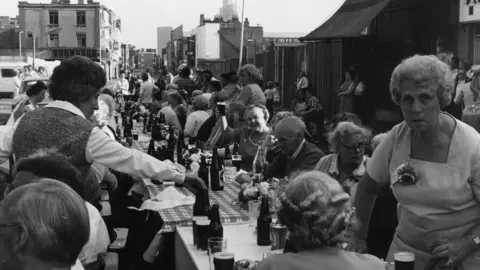 Getty Images People gathered around a long table set with bottles and food for a street party for the Queen's Jubilee in 1977