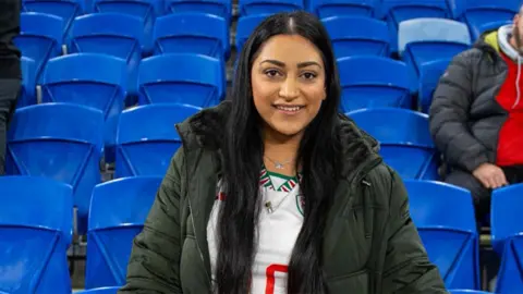 Roopa Vyas Roopa, a South Asian woman smiling. She is wearing a white football top with a green jacket on top. There are blue stadium seats behind her.