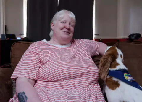 BBC Kayleigh at home on the sofa with dog Rosie next to her. Kayleigh has blond hair and is wearing a pink striped top. She is smiling towards the camera