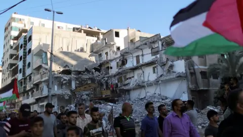 Reuters Palestinians hold a Palestinian flag and walk past a building destroyed by Israeli air strikes in the northern Gaza Strip as they take part in a protest at the Jerusalem Day flag march (15 June 2021)