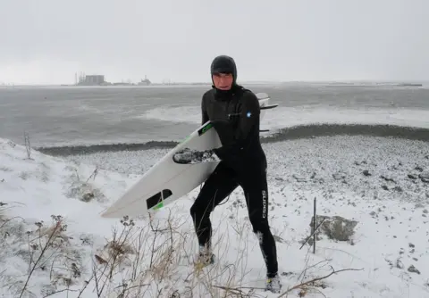 Getty Images Surfer holding board walks up slope amid a snowy landscape