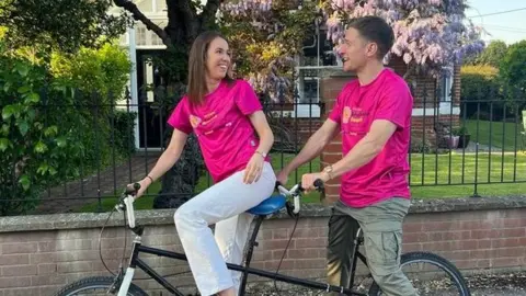 Brain Tumour Research/Handout Ms Lowe and Mr Walklin wearing pink tshirts and sitting on a tandem bike