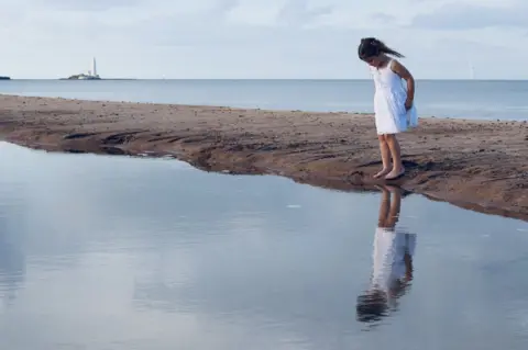 Louise Warburton A girl looks at her reflection in a calm lagoon next to the sea