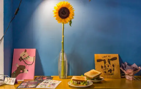 BBC A drawing of a cow flipping off visitors with the message 'leave my tits alone', next to a sunflower in a vase on a table against a blue wall