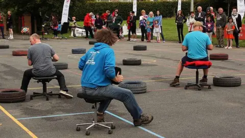 World Alternative Games Three men race around a course on office chairs watched by a crowd