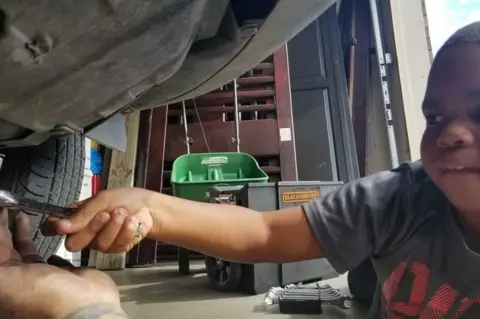 Adam Ely Tysen reaches out underneath his mum's car, with a helping hand from Adam