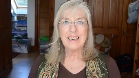 Diana Garland Diana, a woman smiling at the camera. She has a patterned scarf around her neck and is wearing a brown top. The background is the inside of a house, with brown wooden doors, and an open door.