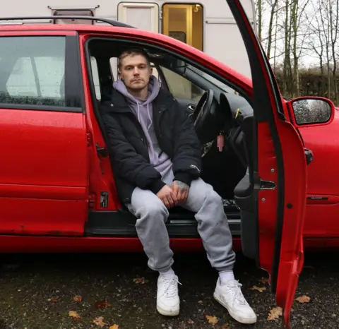 A young man sits in the driver's seat of a red saloon car, with the door open and him facing outwards, his white trainers brushing the floor. He's got short, blonde hair and stubble, a black flesh tunnel piercing in his right earlobe, and an industrial bar piercing - also black - across the top of his ear. He wears a black jacket over a lilac hoodie and grey joggers. In the background behind the car a caravan is visible, the door slightly open and warm yellow light coming from inside.