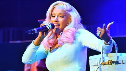 Rapper Cardi B performing on stage. She has bleach blonde hair with is dyed pink at the ends where it has been curled. She is wearing a turquoise long-sleeved dress and is holding a microphone to her mouth with her right hand while she sings/raps. She also has a turquoise handbag hanging from her left arm with the straps around her wrist and her nails are painted turquoise to match her dress.