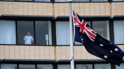 An Australian flag flies in front of a hotel building where a man looks out from his quarantine room