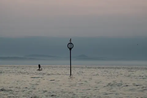 Joanne Baird At the End of the Day - a paddleboarder at sea, next to a metal structure sticking out of the water