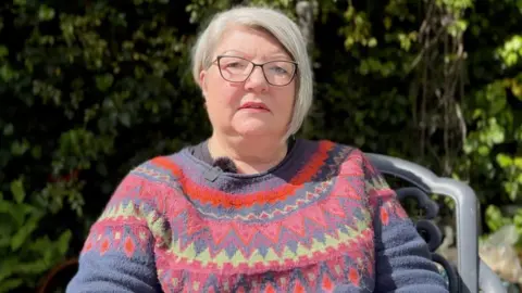 Christine Morgan with blond hair wearing a patterned jumper sitting outside