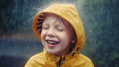 Getty/Sally Anscombe Little boy in raincoat smiles as it rains