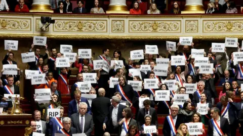 Reuters Supporters of the no-confidence motions hold placards in the National Assembly reading 'retrait', '64 ans c'est non' and 'on continue'