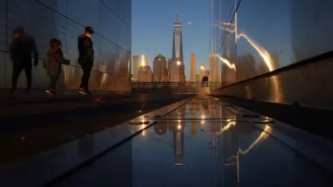 Getty Images People walk through the Empty Sky 9/11 Memorial as the moon rises next to One World Trade Center in New York City at sunset on February 25, 2021 as seen from Jersey City, New Jersey