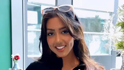 Sanam Harrinanan A head and shoulders photo of Sanam Harrinanan smiling for the camera in a cafe. She has a pair of sunglasses on the top of her head and her brunette hair is worn down. She is wearing a black top and behind her is a window. You can just make out a small vase of flowers on another table and a fern plant to the right-hand side of her head