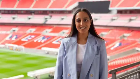 Hina Shafi Hina Shafi wearing a light blue blazer and stood smiling inside Wembley Stadium which is behind her