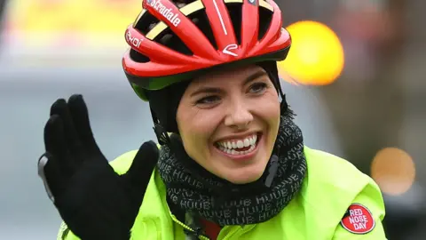 A woman rides a bicycle on a rain-covered road with an escort car trailling behind. She's smiling and waving, and wears a bright red crash helmet and a bright yellow hi-vis jacket.