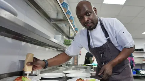 Anti Banquet Chef George Opondo looks at the camera as he applies garnishes to dinner plates in a large kitchen