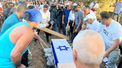 Laurie Margolis A coffin is lowered into a grave watched on by mourners