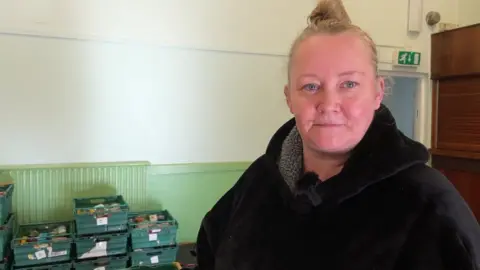 Still of Donna Hayhow in a black fleece in front of donations at the foodbank.