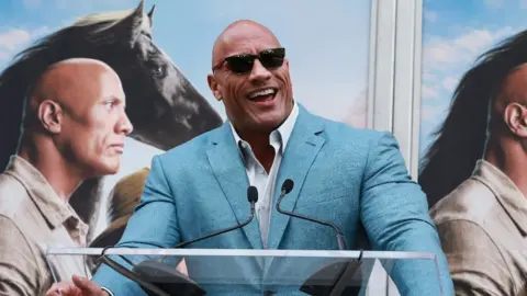 Getty Images The Rock speaks at a podium during his latest film release