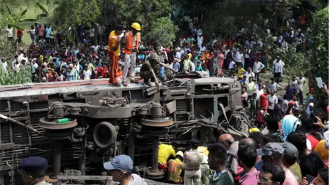 EPA Dozens of local people gathering near an overturned train carriage, with three emergency service workers standing on top of it