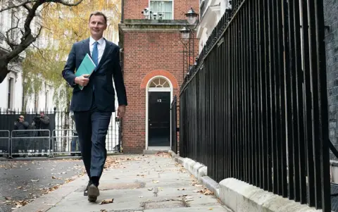 PA Media Jeremy Hunt walking outside number 11 Downing Street in a suit and blue tie holding a green budget statement in November 2023