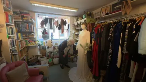 Mrs Ellicott bending down near the wedding dress which is displayed in a small shop