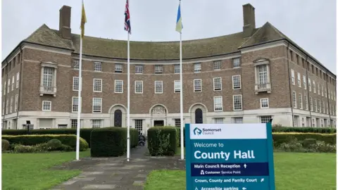 Daniel Mumby The exterior of Somerset County Hall with three flagpoles and a teal-coloured sign