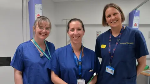 NHS Somerset Foundation Trust Jo Morrison, Dr Holly Baker-Rand and Dr Becky Newhouse smile at the camera while wearing medical scrubs