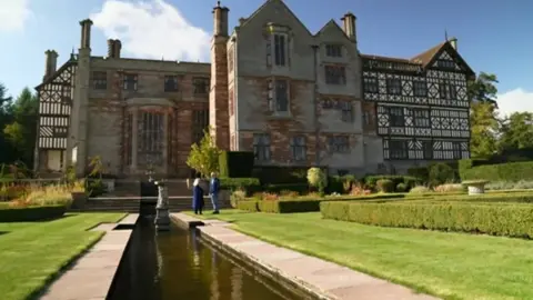 Two people in the distance standing in front of a mansion by a large pond