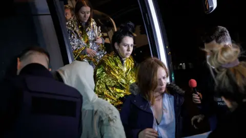 Getty Images Two women wrapped in metal blankets walk off a bus