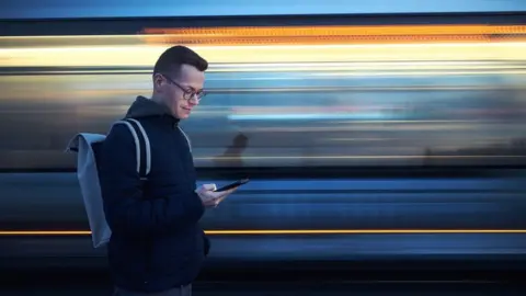 Jaromir/Getty Images Man with backpack holding and using phone against train in blurred motion - stock photo