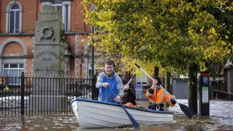 PA Media Three men row a boat along a flooded street in Newry city centre