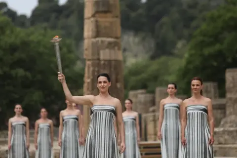 George Vitsaras/EPA-EFE Greek actress Mary Mina (centre), in the role of the High Priestess, raises the torch of the Olympic Flame during the Olympic Flame lighting ceremony