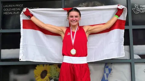 Elise Glynn Elise Glynn, a white, brunette woman, wears a red boxing uniform and holds an England flag above her head. She is smiling and wearing a medal around her neck