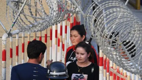 Getty Images A file photo from 2019 showing Uygher women going through the entrance to a bazaar in Hotan, Xinjiang, past police officers and a gateway covered in razor wire